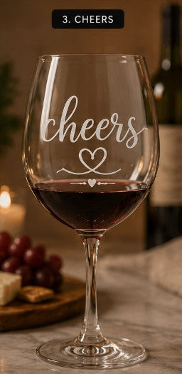 Wine glass with 'cheers' and a heart design on a table with grapes and cheese.