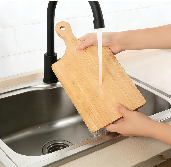 Person washing a wooden cutting board under running water in a kitchen sink.